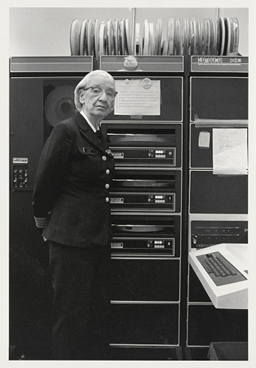 A person in military uniform, glasses, and short white hair stands in front of a classic tape-operated computer.